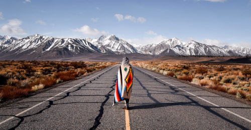 Person wearing Native American blanket, walking down highway towards mountains