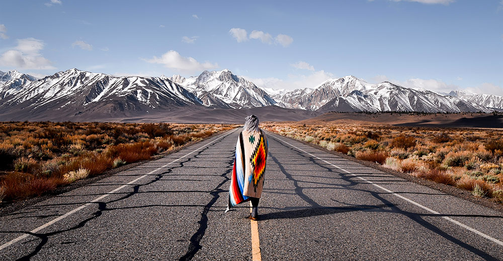 Person wearing Native American blanket, walking down highway towards mountains
